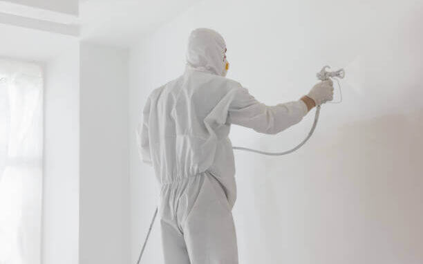 Decorator Painting White Wall With Spray Indoors, Back View